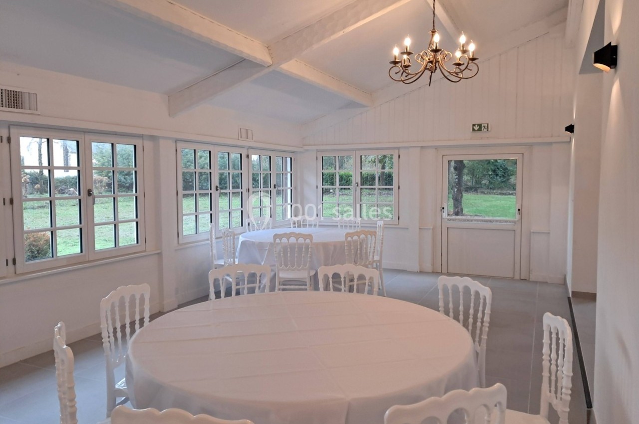 Salle lumineuse avec tables rondes dressées, chaises blanches et grandes fenêtres donnant sur un jardin verdoyant.