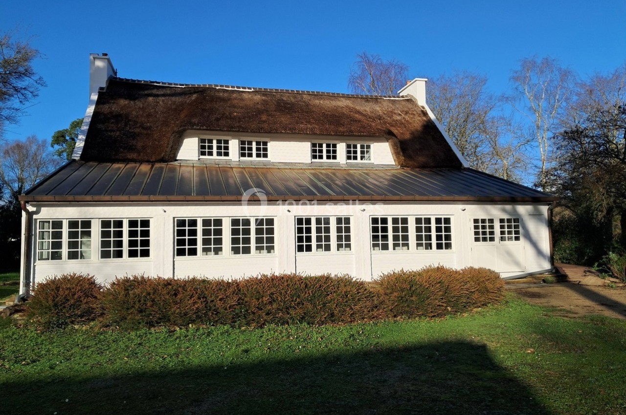 Maison blanche avec toit de chaume, grandes fenêtres et jardin, éclairée par le soleil sous un ciel bleu.