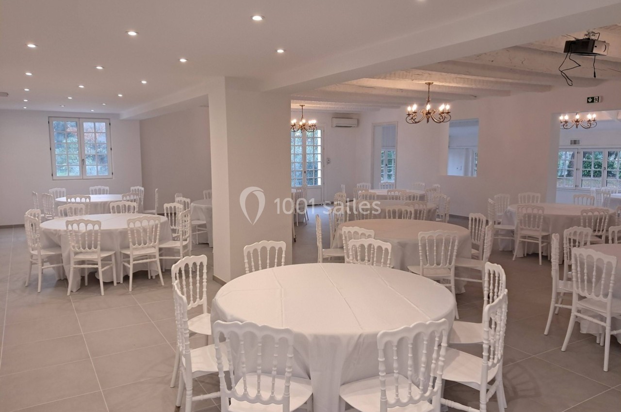 Salle lumineuse avec tables rondes dressées de nappes blanches et chaises blanches, éclairée par des lustres et spots.