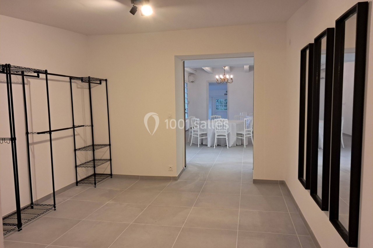 Pièce vide avec étagères métalliques à gauche, miroirs à droite et vue sur une salle avec des chaises blanches au fond.