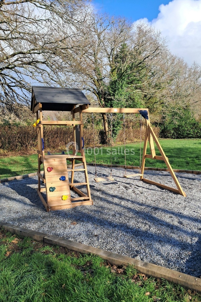 Structure de jeux en bois avec toboggan, mur d'escalade et balançoires, installée sur une surface de gravier dans un parc.