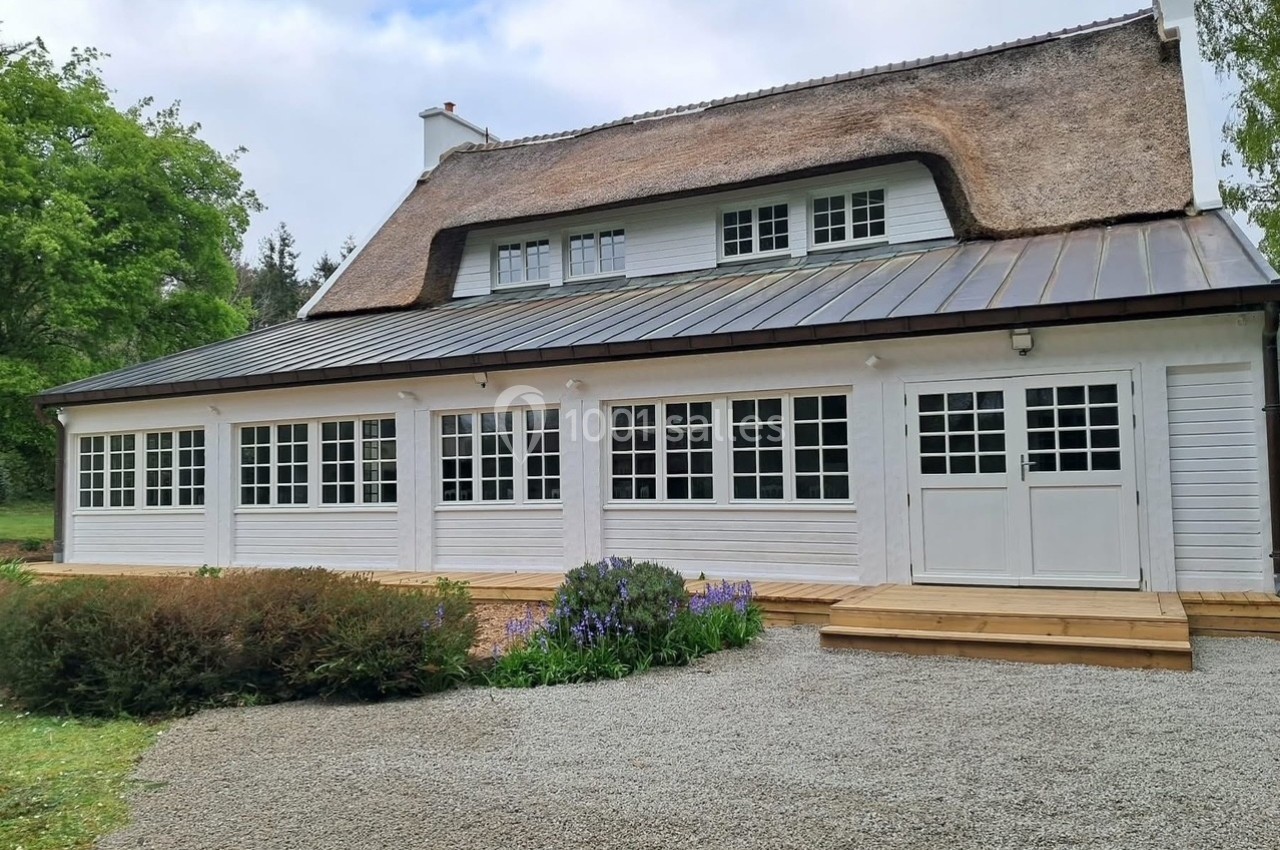 Maison blanche avec toit de chaume, grandes fenêtres, terrasse en bois, entourée de végétation et d'une allée gravillonnée.