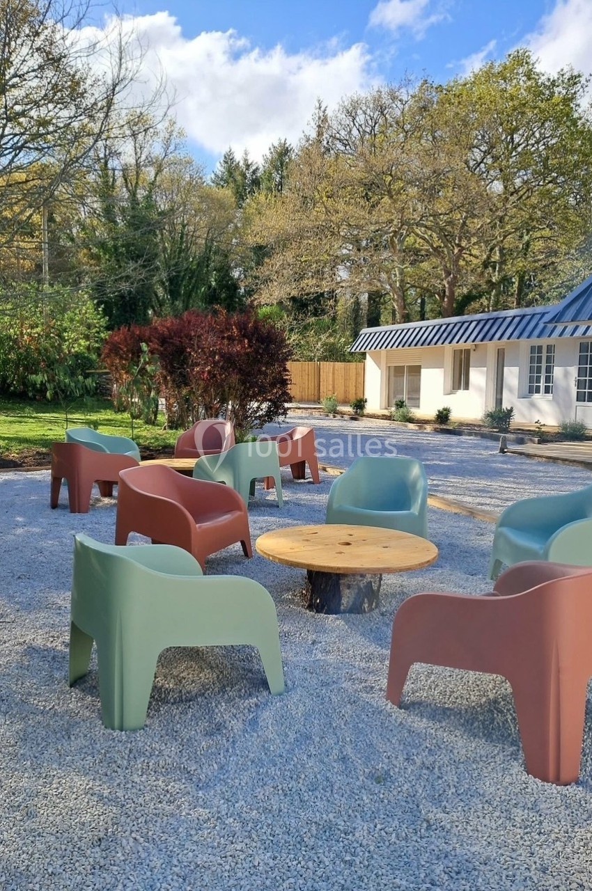 Chaises colorées et table ronde en bois disposées sur un sol gravillonné devant un bâtiment blanc entouré d'arbres.