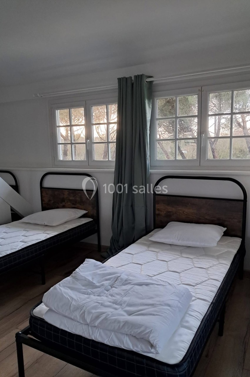 Deux lits simples avec draps blancs dans une chambre lumineuse, fenêtre avec rideaux verts et vue sur des arbres.