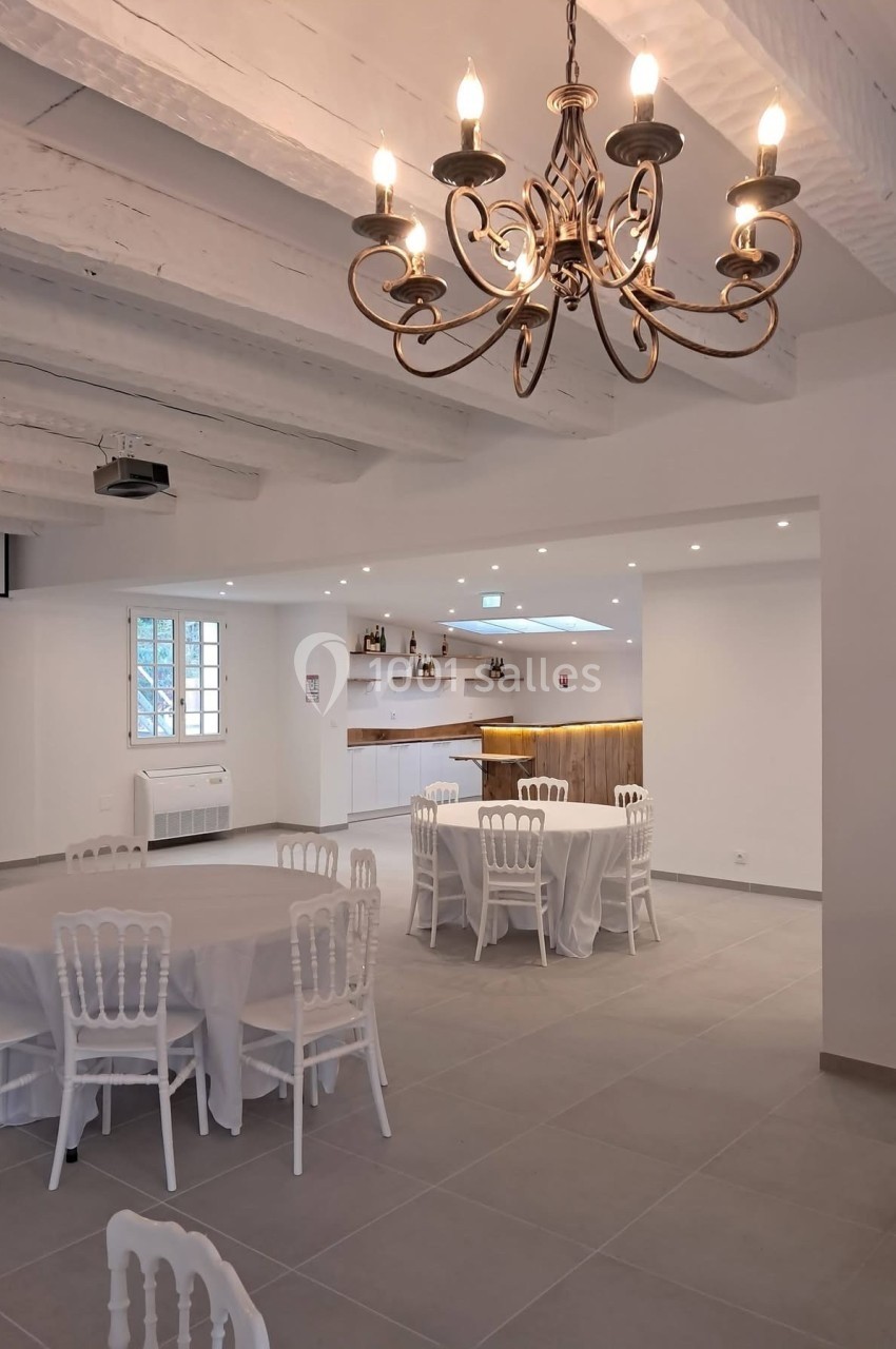 Salle lumineuse avec tables rondes dressées de nappes blanches, chaises assorties et lustre suspendu au plafond en poutres…