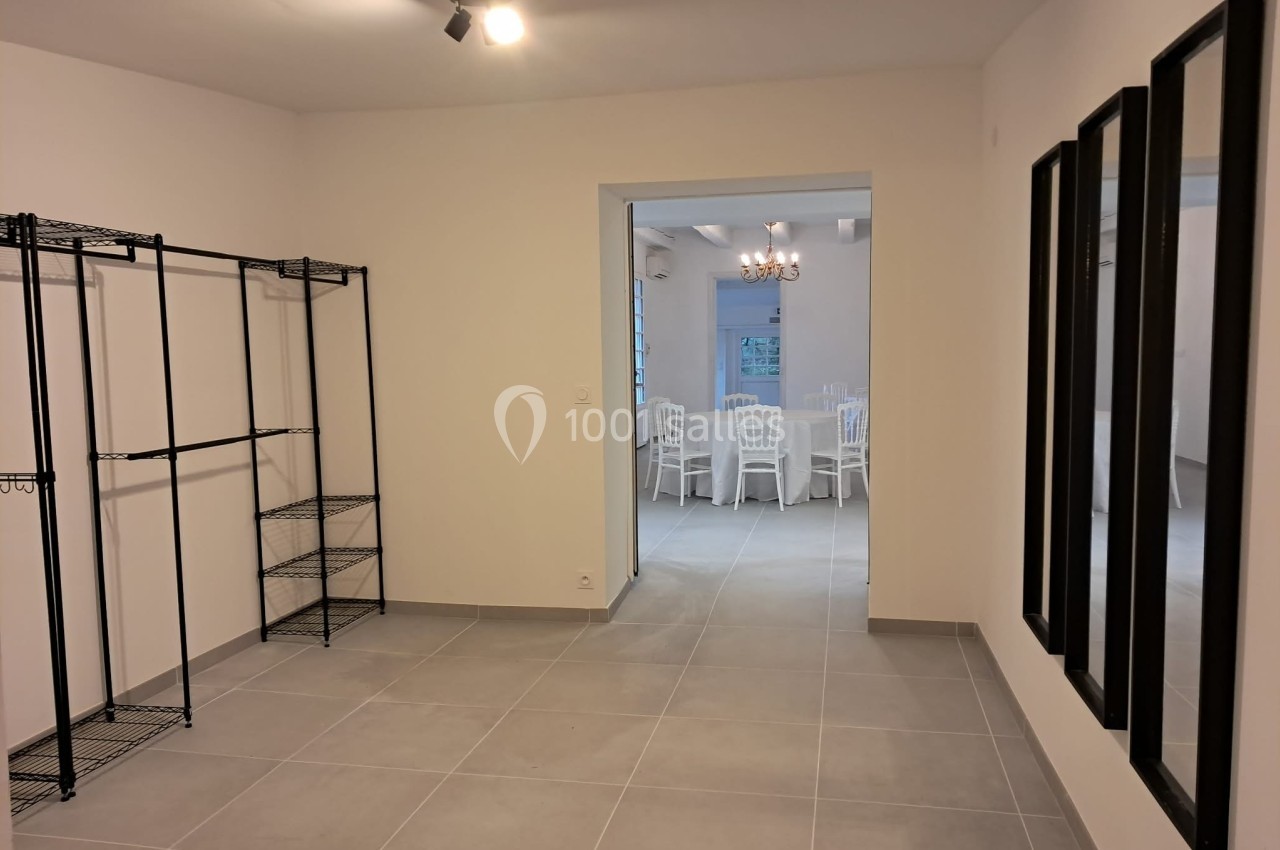 Pièce vide avec étagères métalliques à gauche, miroirs à droite et vue sur une salle avec des chaises blanches au fond.