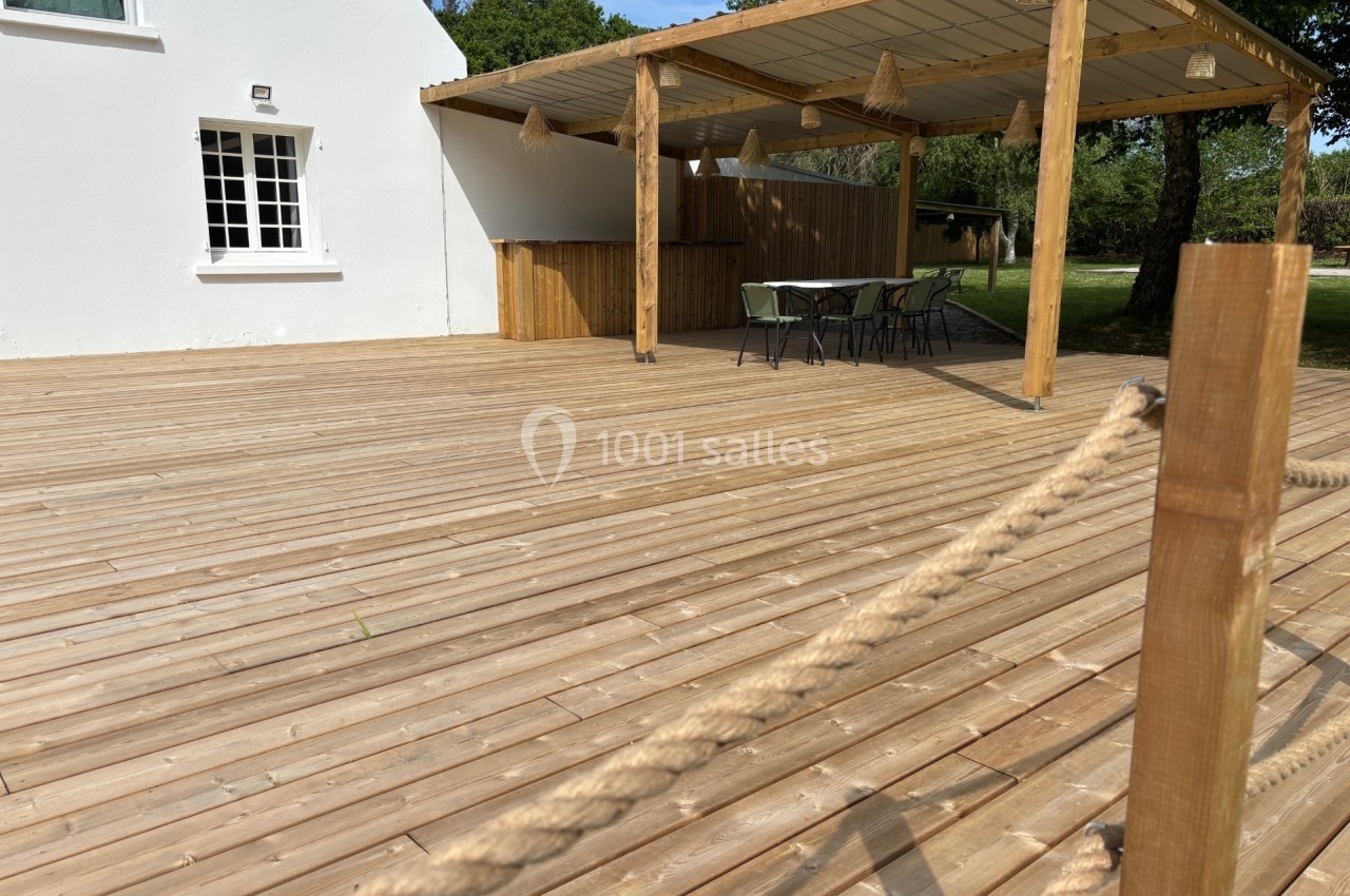 Terrasse en bois avec pergola, table et chaises, entourée de verdure et attenante à une maison blanche.