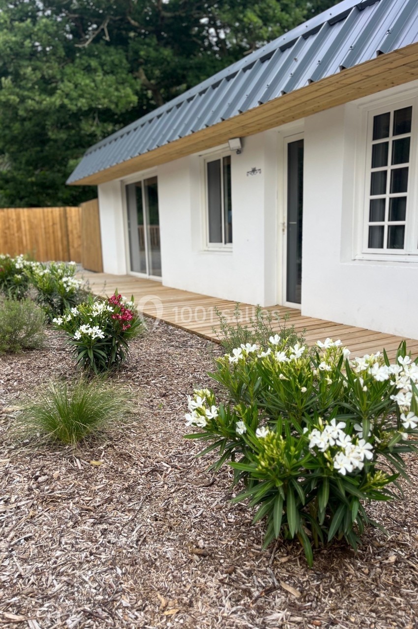 Façade d'une maison blanche avec toit en métal, terrasse en bois et parterre de fleurs devant une clôture en bois.
