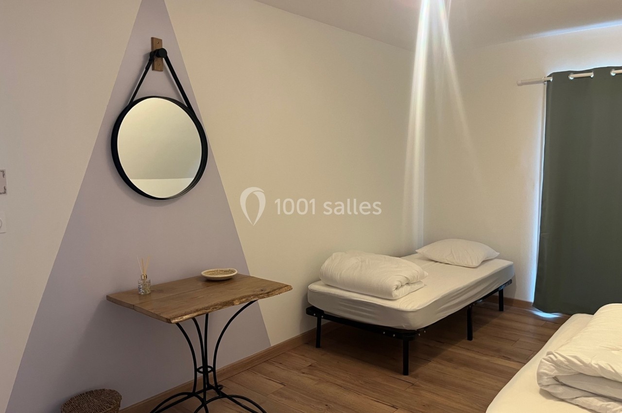 Chambre simple avec deux lits, une table en bois, un miroir mural rond et un rideau vert couvrant une fenêtre.