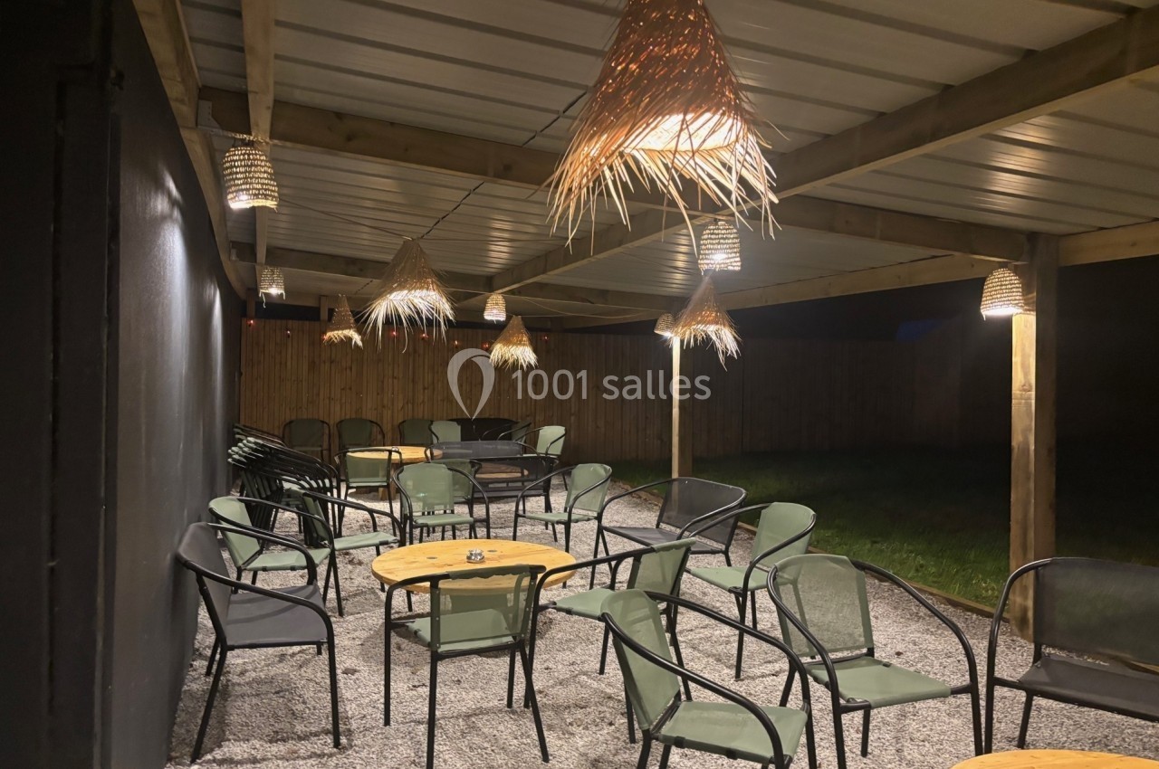 Terrasse couverte éclairée de nuit, avec des tables rondes en bois et des chaises noires sur un sol en gravier.
