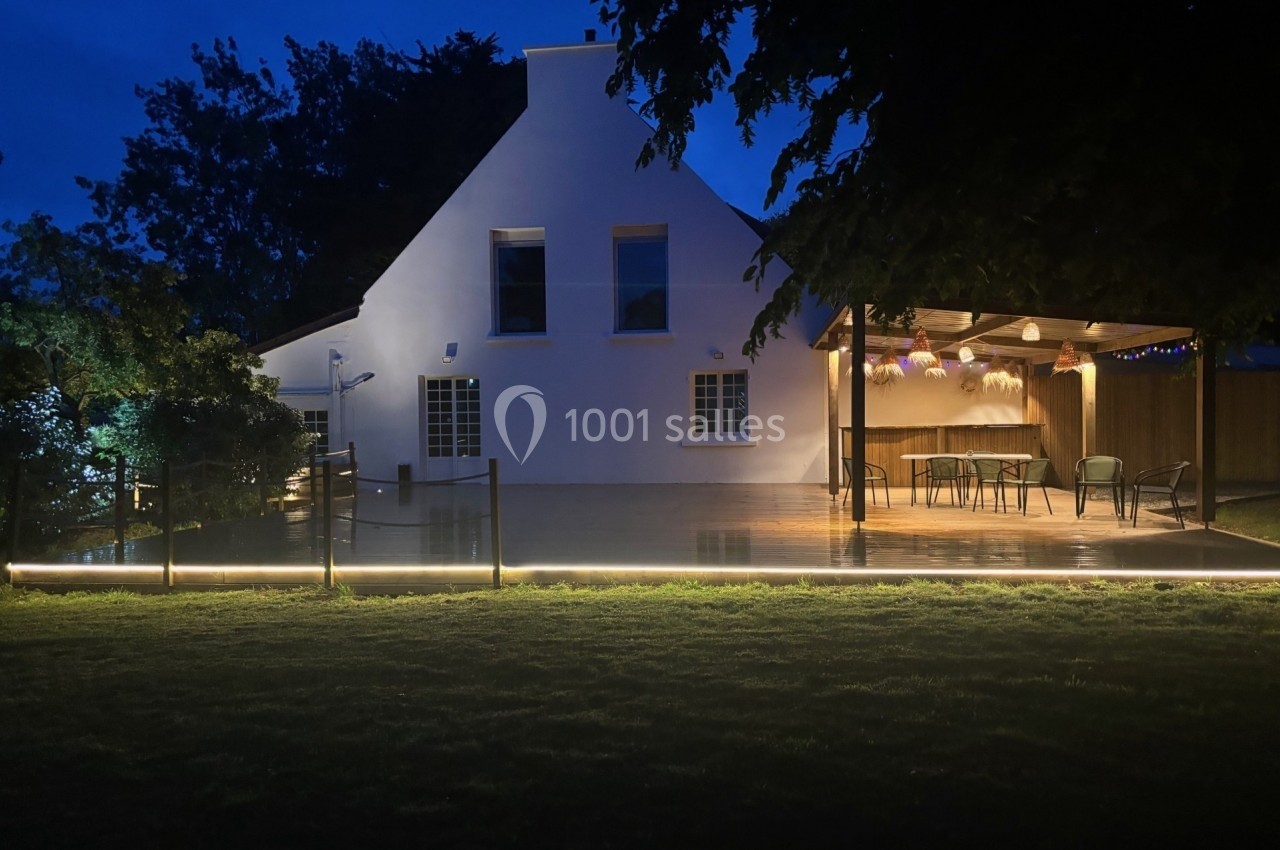 Maison blanche éclairée la nuit, terrasse en bois illuminée avec pergola et mobilier de jardin, entourée de verdure.
