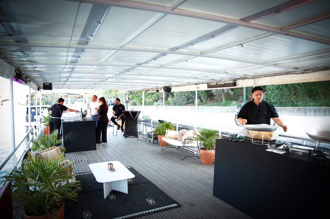 Terrasse couverte avec personnel préparant un buffet, entourée de plantes et offrant une vue sur l'extérieur.