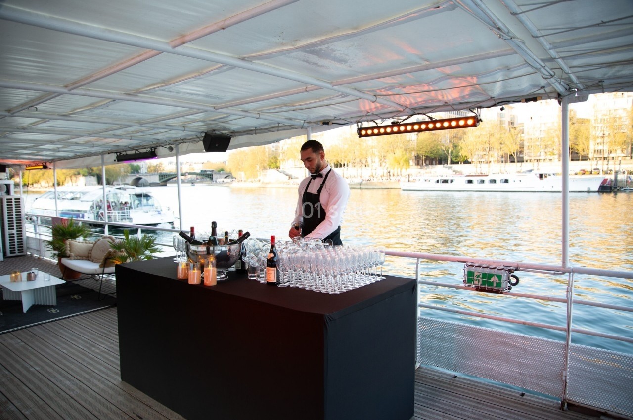 Serveur préparant un bar avec des verres et des bouteilles sur une péniche amarrée près d'une rivière.