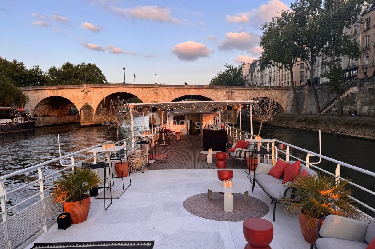 Terrasse d'une péniche aménagée avec des sièges et des plantes, amarrée près d'un pont sur une rivière au coucher du soleil.