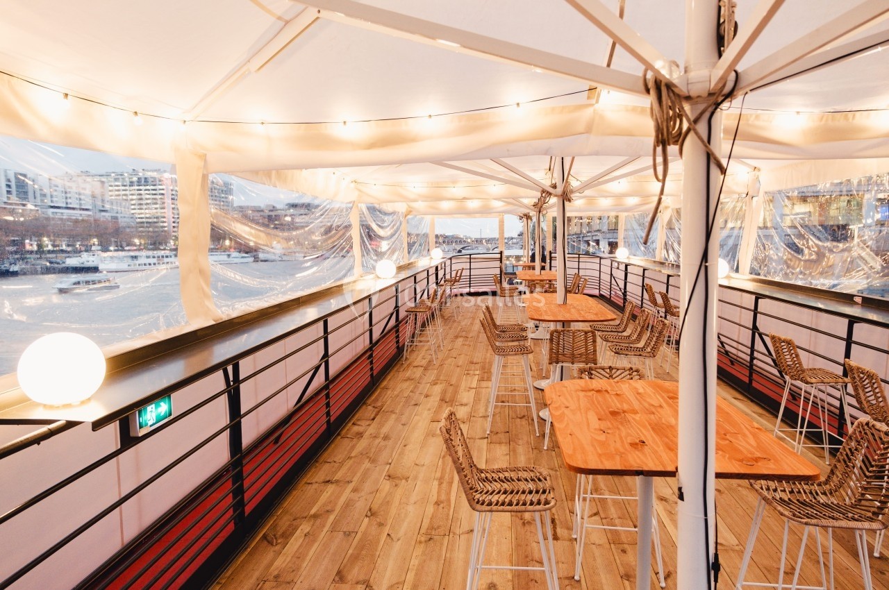 Terrasse couverte d'un bateau avec tables et chaises en rotin, éclairée par des guirlandes lumineuses au bord d'une rivière.