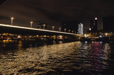 Vue nocturne d'un pont illuminé au-dessus d'une rivière, avec des bâtiments en arrière-plan et des reflets sur l'eau.