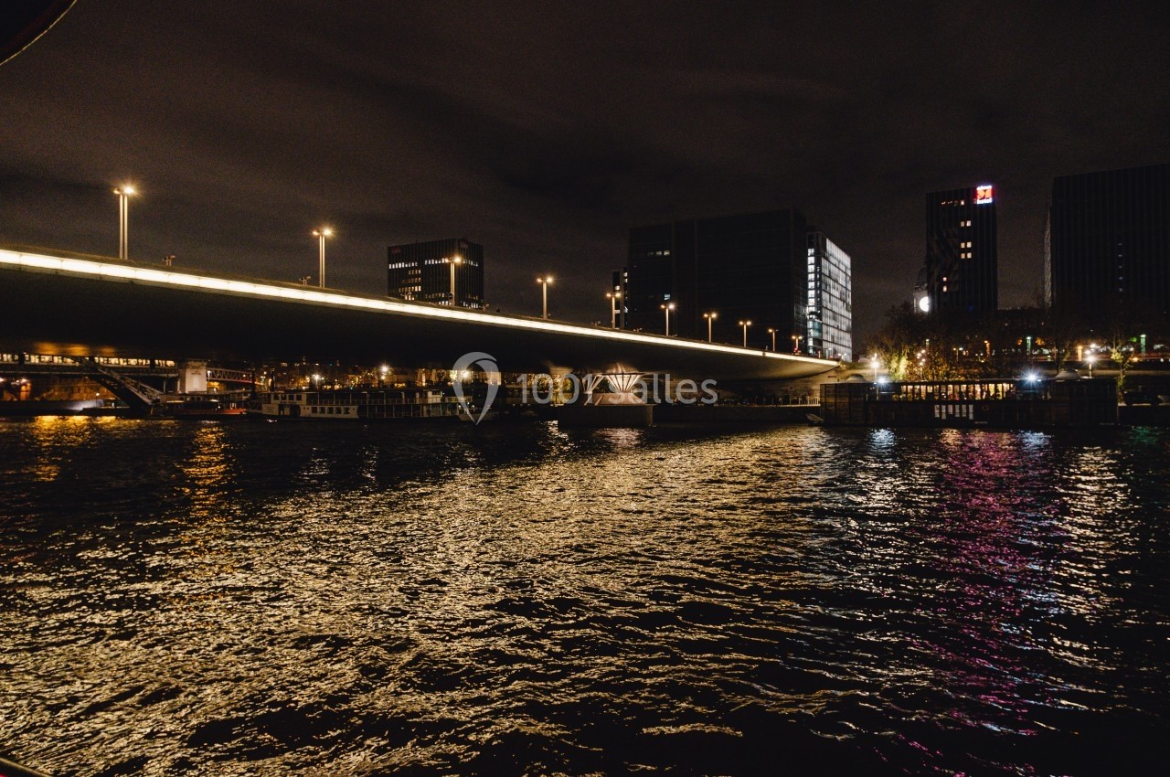 Vue nocturne d'un pont illuminé au-dessus d'une rivière, avec des bâtiments en arrière-plan et des reflets sur l'eau.