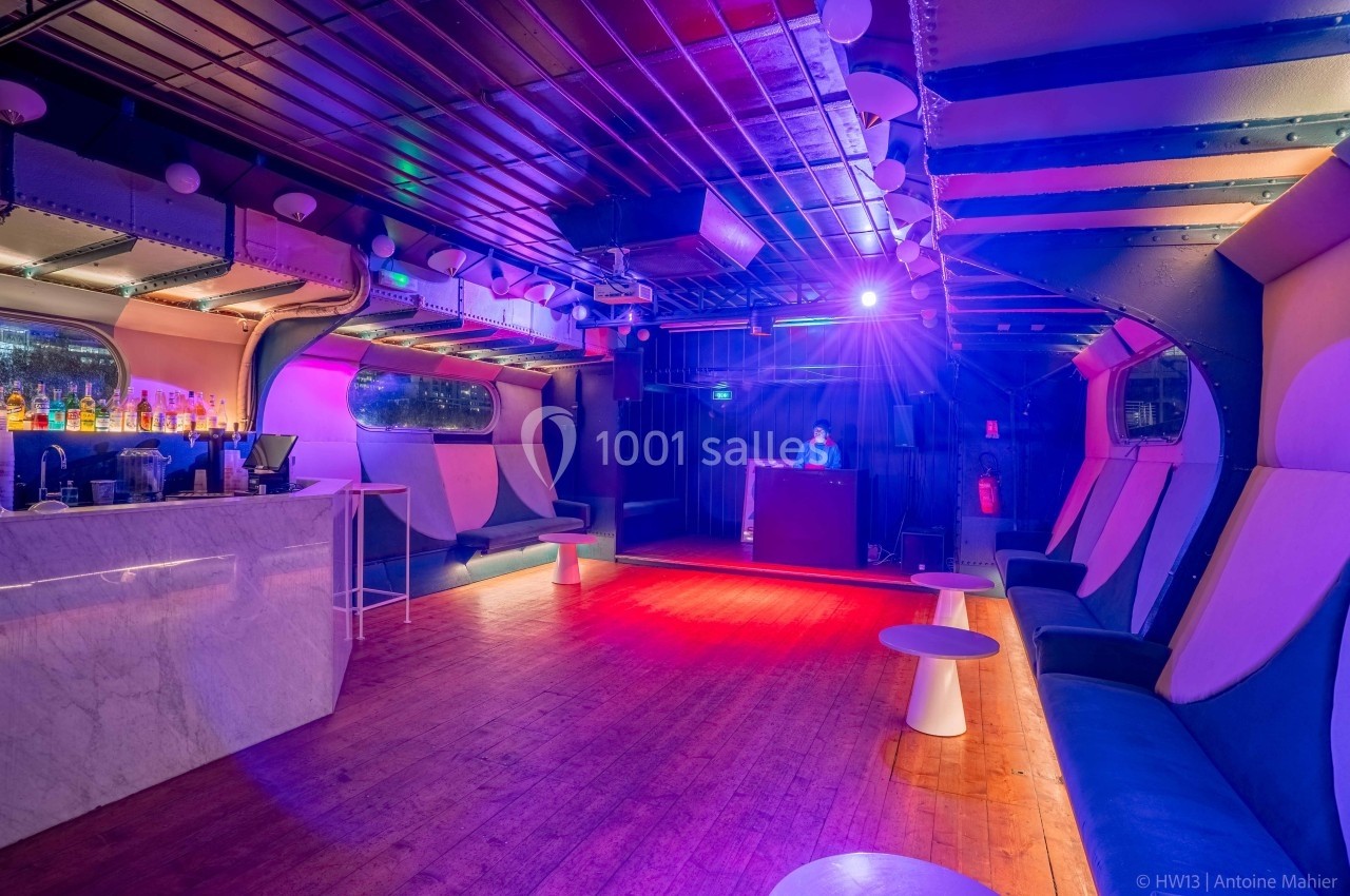 Salle de bar avec éclairage coloré, banquettes, tables basses et un DJ en arrière-plan.
