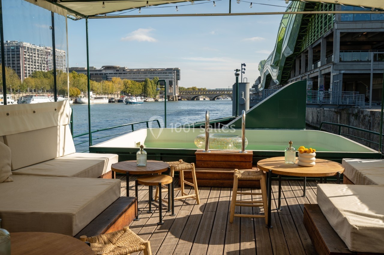 Terrasse en bois avec canapés, tables et vue sur la Seine, entourée de bâtiments modernes et d'un pont.