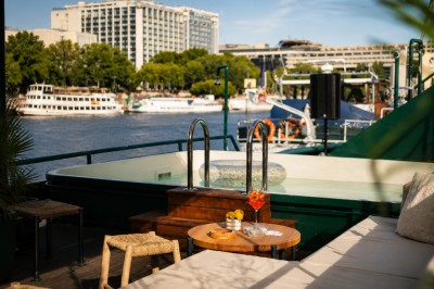 Terrasse aménagée sur une péniche avec vue sur la Seine, des bâtiments et un pont en arrière-plan.