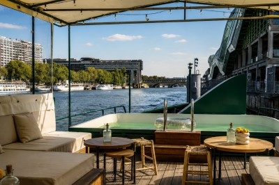 Terrasse aménagée sur une péniche avec vue sur la Seine, des bâtiments et un pont en arrière-plan.