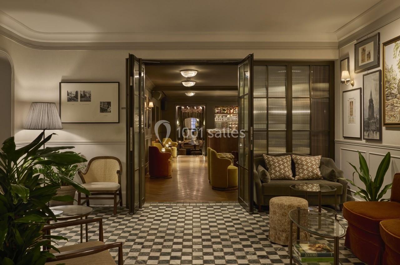Salon élégant avec sol en carreaux noirs et blancs, fauteuils confortables, plantes et vue sur un bar éclairé.