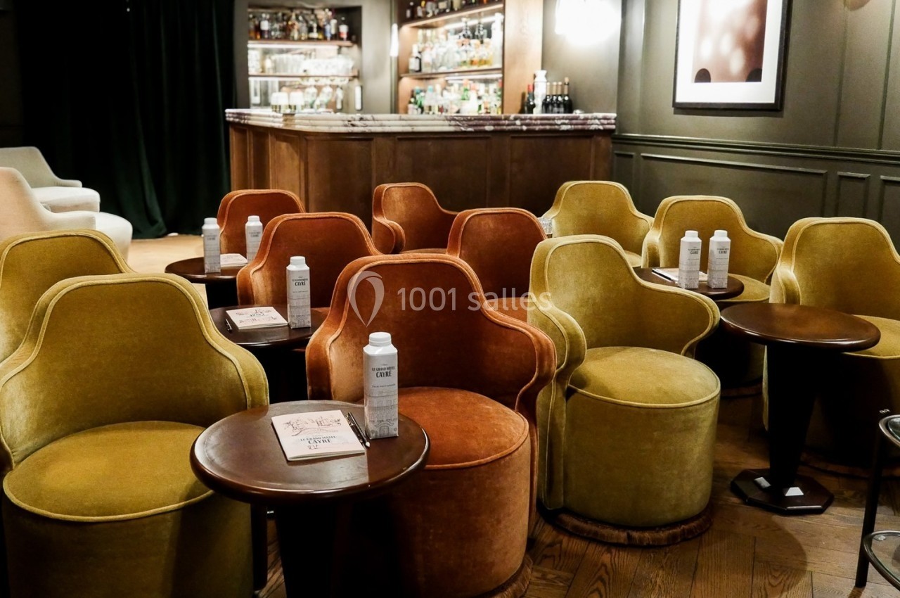 Fauteuils colorés disposés autour de petites tables en bois dans un salon élégant avec un bar en arrière-plan.