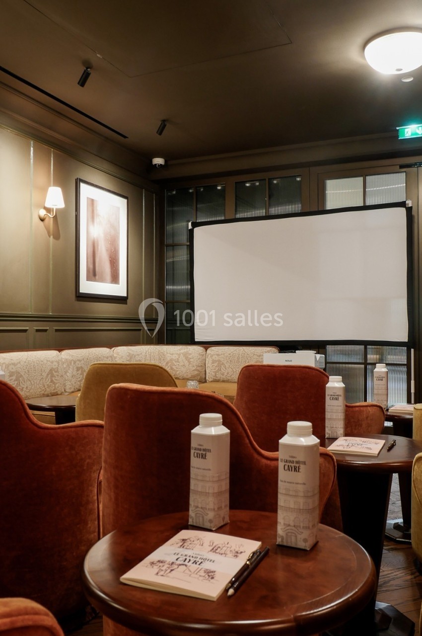 Salle de réunion confortable avec fauteuils en velours, tables en bois, écran de projection et carnets disposés.