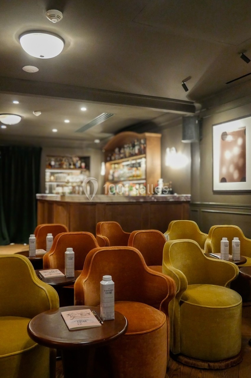 Salle de bar avec fauteuils colorés, tables basses et un comptoir en bois garni de bouteilles à l'arrière-plan.