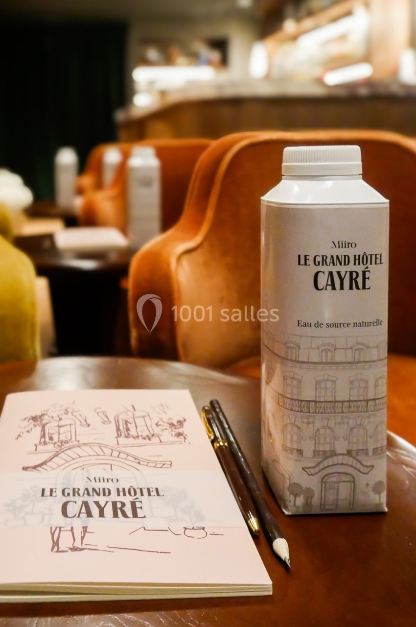 Carton d'eau marqué ’Le Grand Hôtel Cayré’ posé sur une table avec un carnet et des crayons, dans un salon élégant.