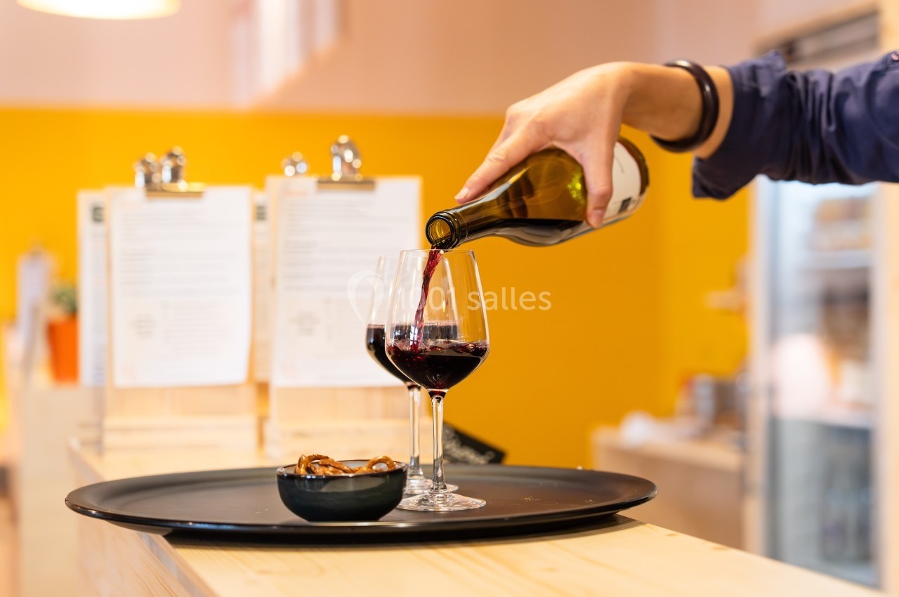 Une main verse du vin rouge dans un verre sur un plateau avec un bol de snacks, dans un intérieur lumineux.
