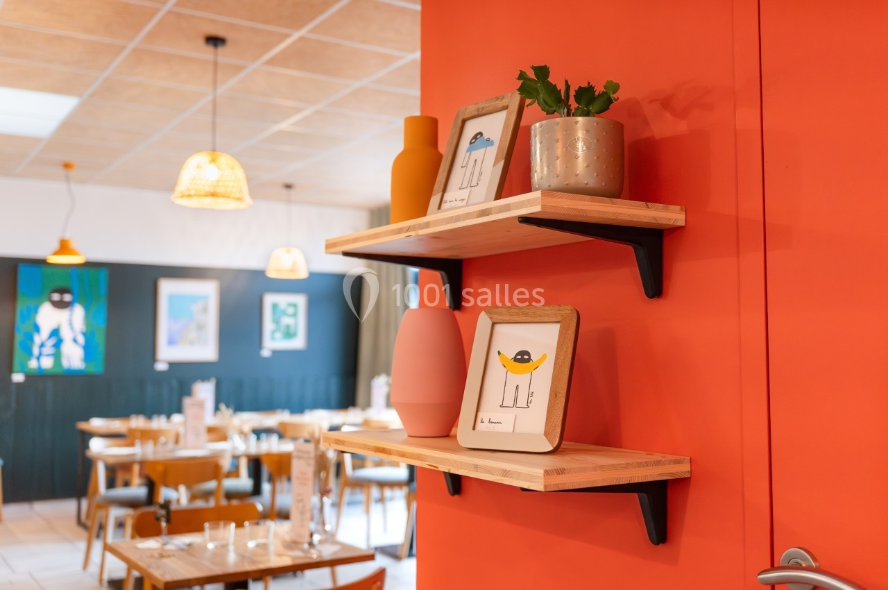 Étagères en bois fixées sur un mur orange, décorées de cadres, vases et une plante en pot, dans un restaurant lumineux.