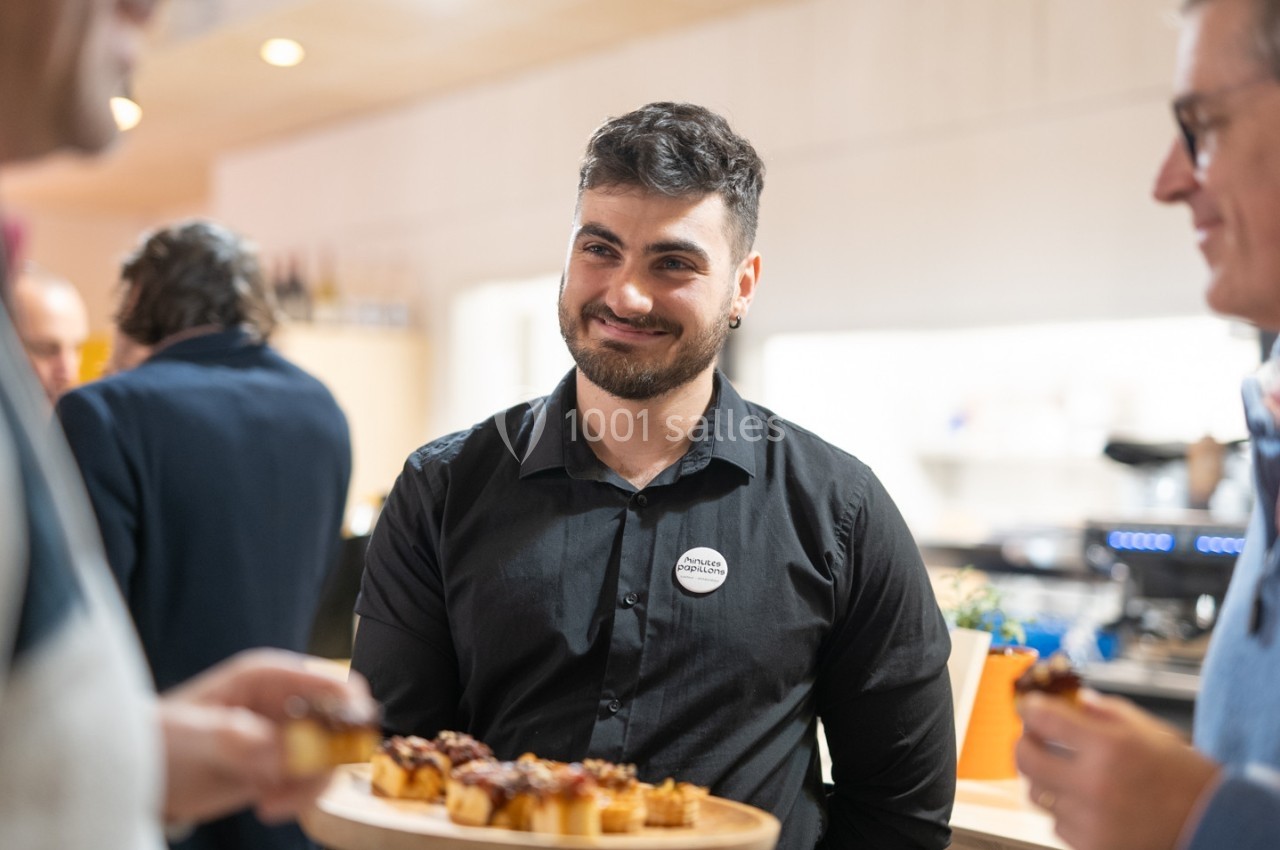 Un serveur souriant en chemise noire propose un plateau de bouchées apéritives dans un espace convivial.
