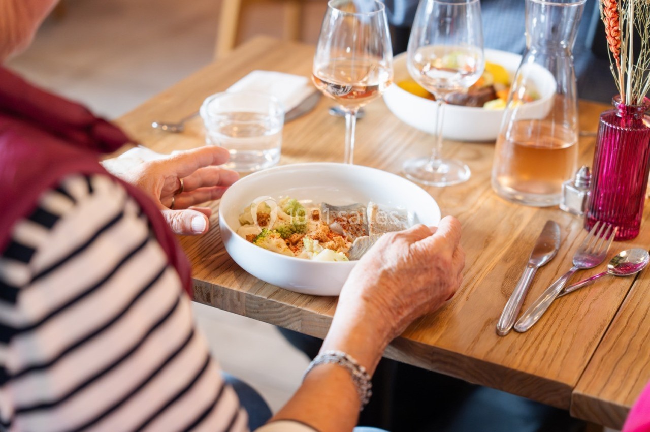 Une personne âgée mange un plat servi dans une assiette blanche, sur une table dressée avec des verres et des couverts.