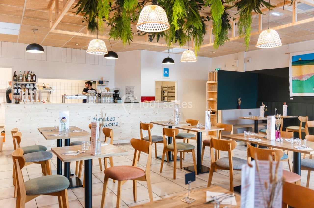 Salle de restaurant lumineuse avec tables en bois, chaises colorées et décoration végétale suspendue.