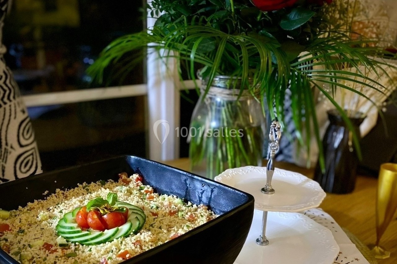 Plat de taboulé garni de tranches de concombre et tomates cerises, posé sur une table décorée avec des fleurs rouges.