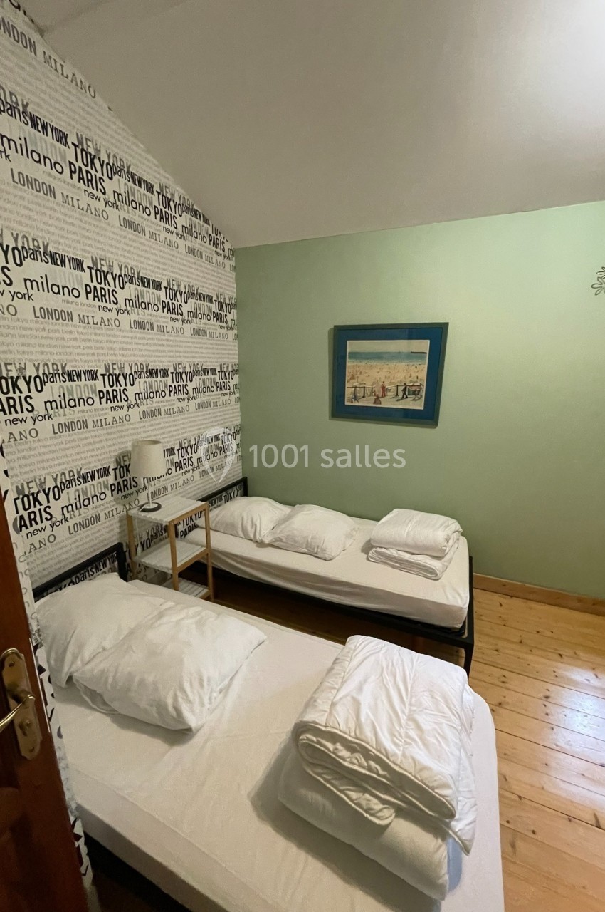 Deux lits simples avec linge de lit blanc dans une chambre au sol en bois, mur décoré de noms de villes.