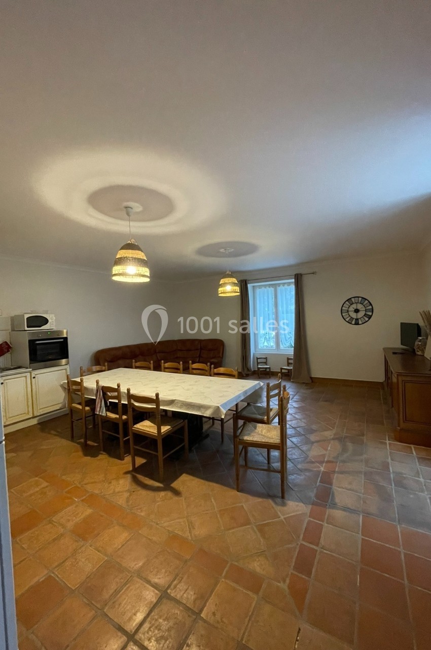 Salle à manger avec une grande table entourée de chaises, cuisine équipée à gauche et salon avec canapé au fond.