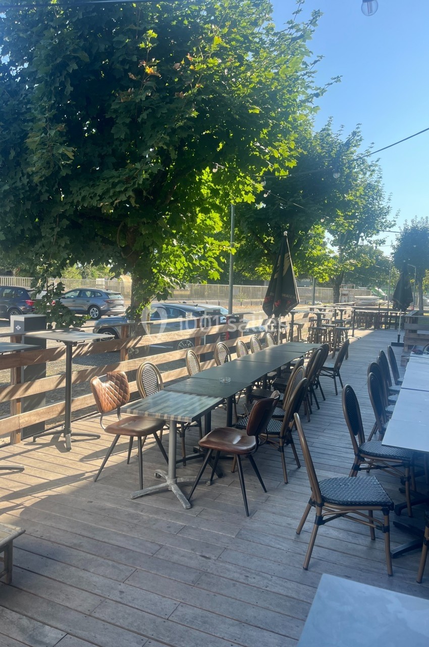 Terrasse en bois avec tables et chaises disposées sous des arbres, bordée par une route et des voitures stationnées.