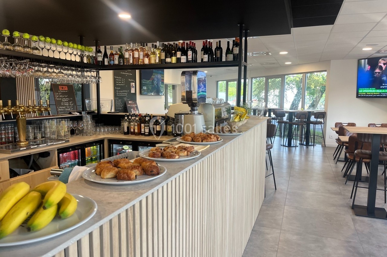 Comptoir d'un café avec viennoiseries, fruits et boissons, entouré de tables et chaises dans un espace lumineux.