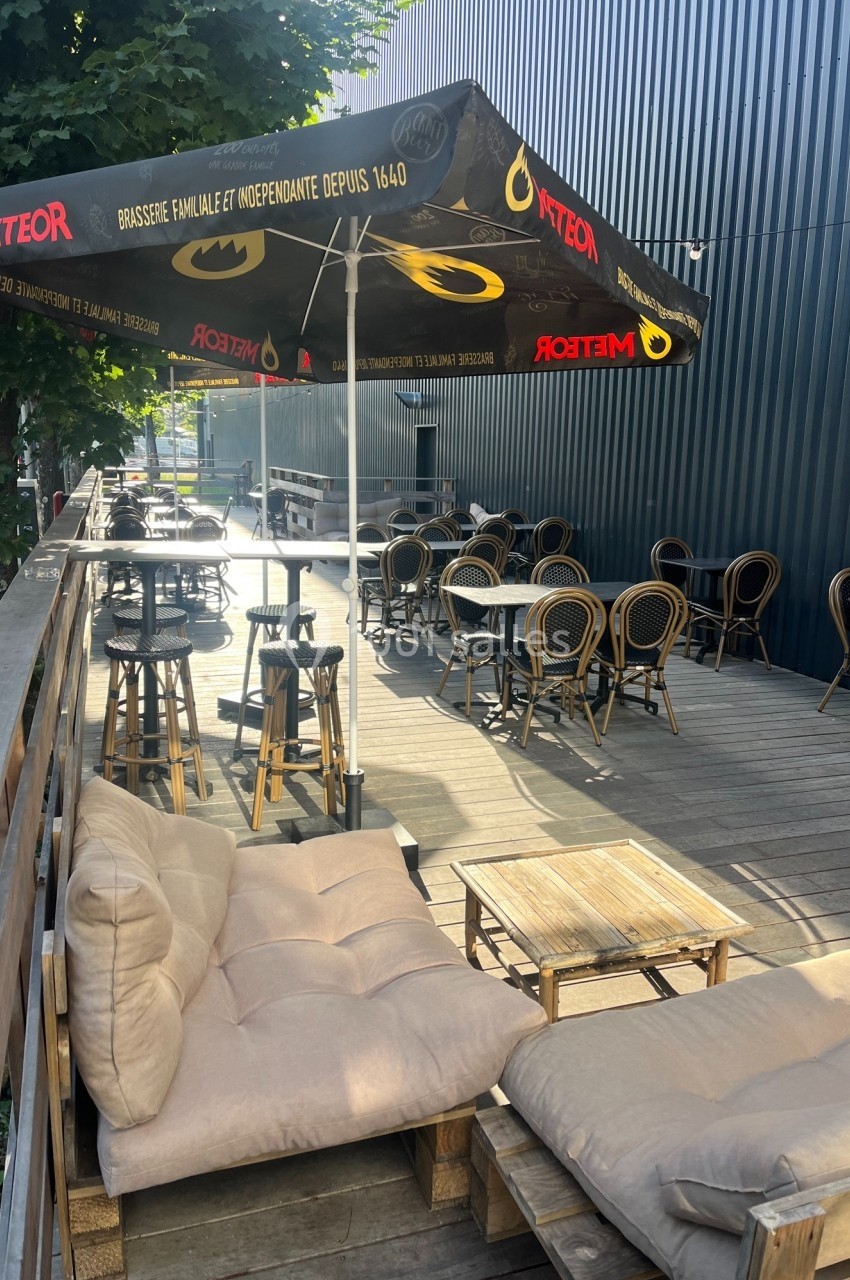 Terrasse en bois avec tables, chaises en rotin et parasols, entourée d'arbres et adossée à un bâtiment sombre.