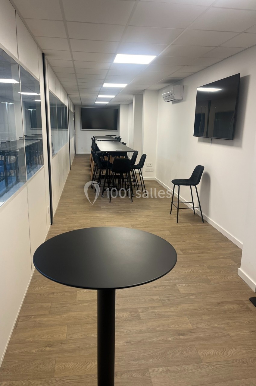 Salle moderne avec longue table, chaises hautes, écrans muraux, sol en bois clair et éclairage au plafond.