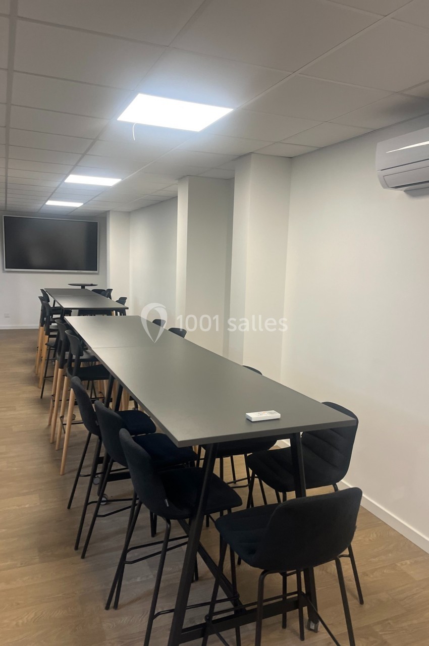 Salle de réunion moderne avec longues tables noires, chaises hautes, écran mural et éclairage au plafond.