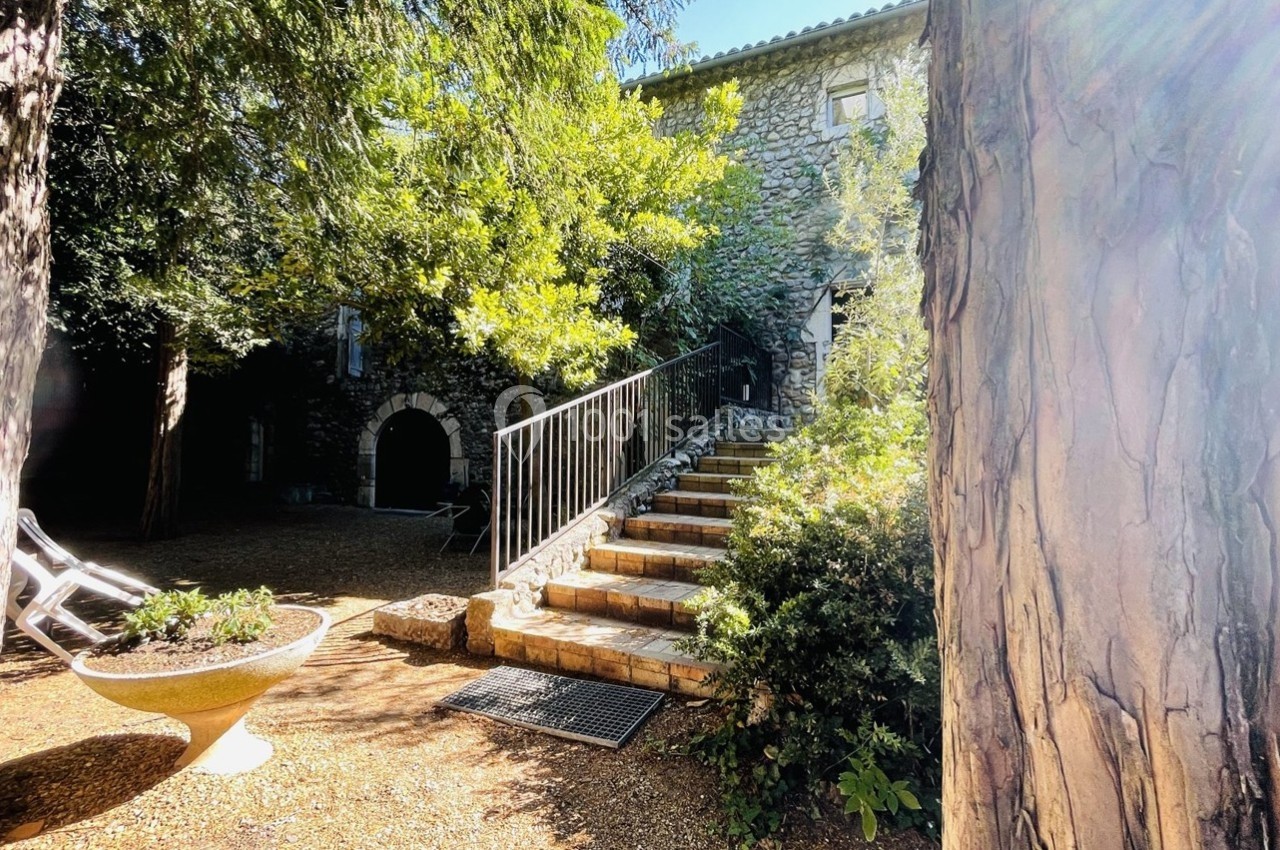 Cour extérieure ensoleillée avec un escalier en pierre menant à une maison en pierre entourée de végétation.