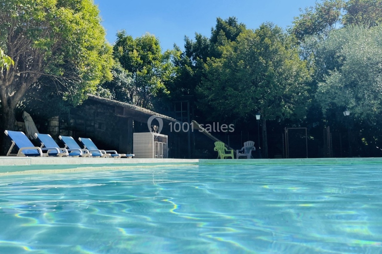 Piscine extérieure entourée de chaises longues bleues, arbres et bâtiment en arrière-plan sous un ciel dégagé.