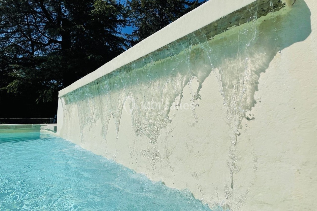 Cascade d'eau claire s'écoulant le long d'un mur blanc dans une piscine extérieure, entourée de végétation.