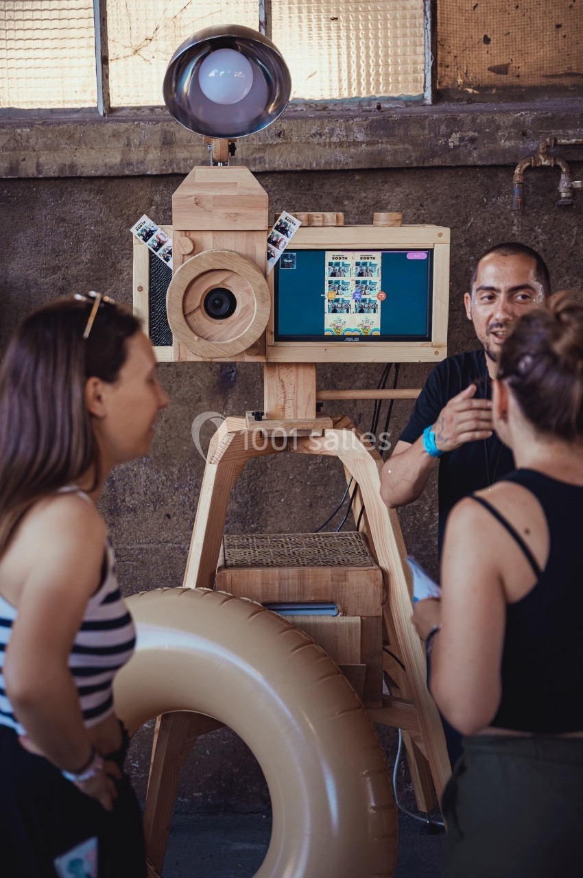Une borne photo en bois avec écran et accessoires, entourée de personnes discutant dans un espace industriel.