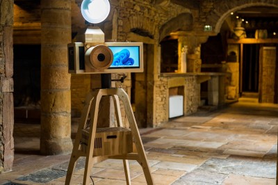Une borne photo en bois avec écran et accessoires, entourée de personnes discutant dans un espace industriel.