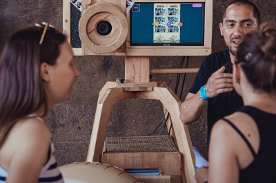 Une borne photo en bois avec écran et accessoires, entourée de personnes discutant dans un espace industriel.