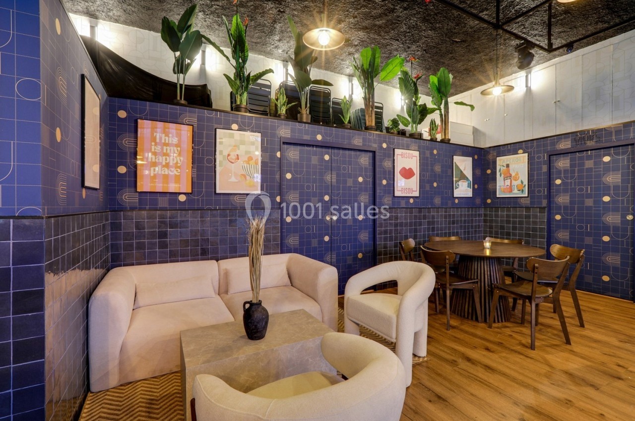 Espace intérieur avec fauteuils, table basse, table à manger en bois, murs bleus décorés et plantes en hauteur.